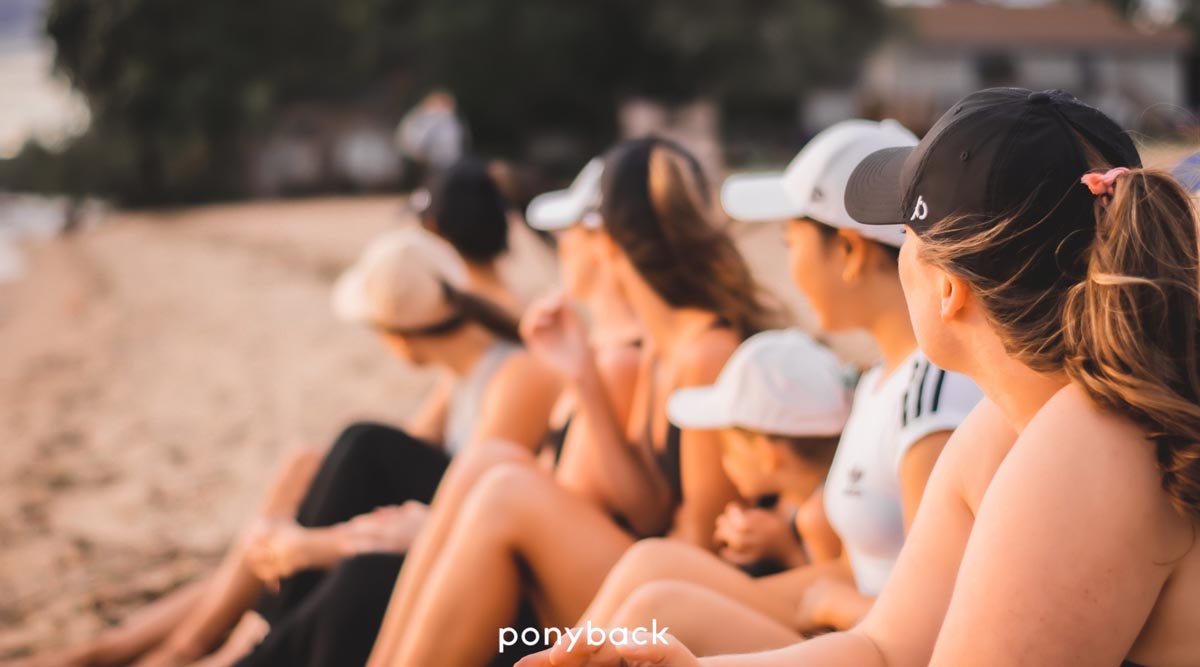 Women wearing ponyback ponytail hats sitting on the beach looking away from the camera, hat colors include white and black and they are all wearing ponytails
