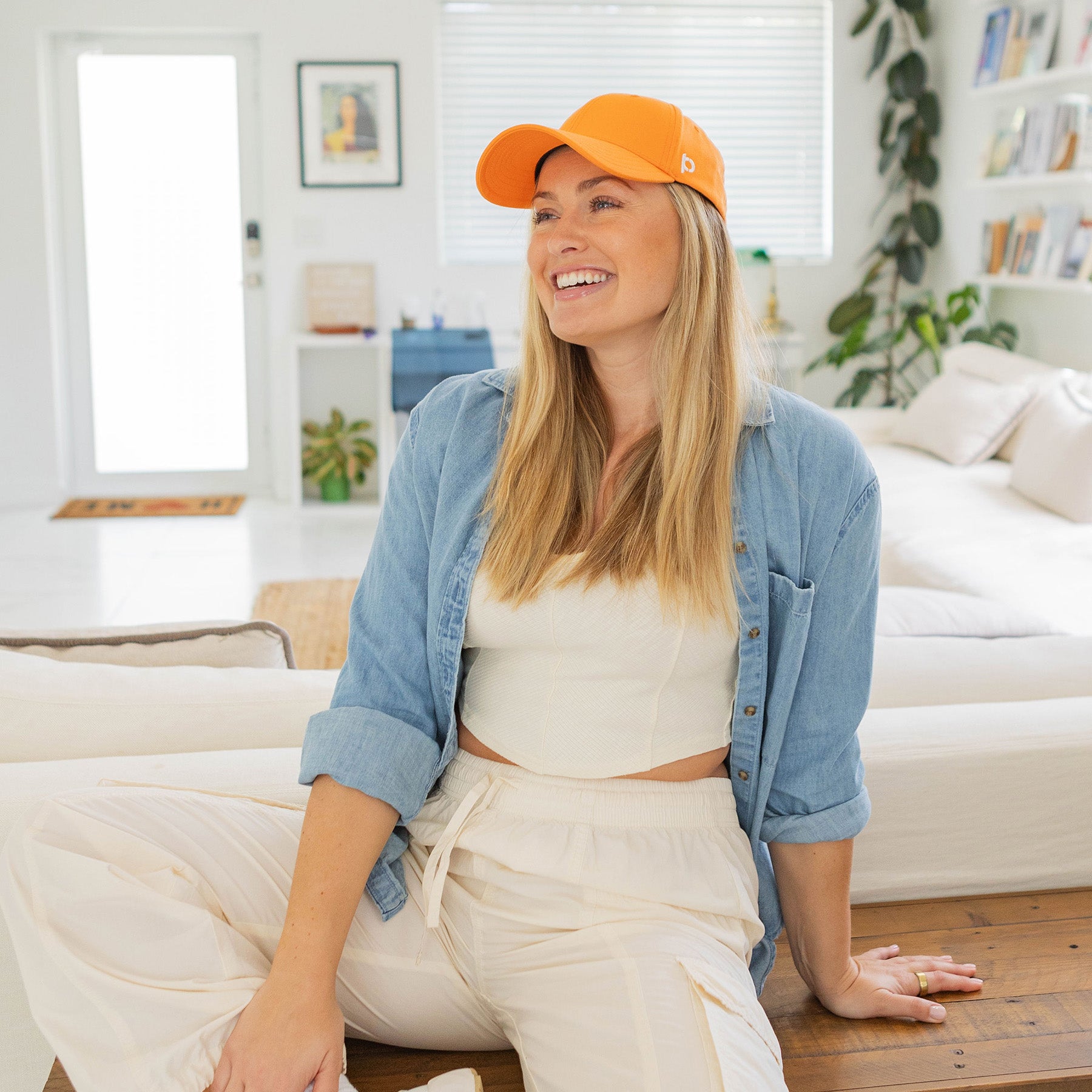 Woman wearing an apricot-colored cap, light blue denim shirt, and white pants, sitting and smiling in a bright, contemporary living room.