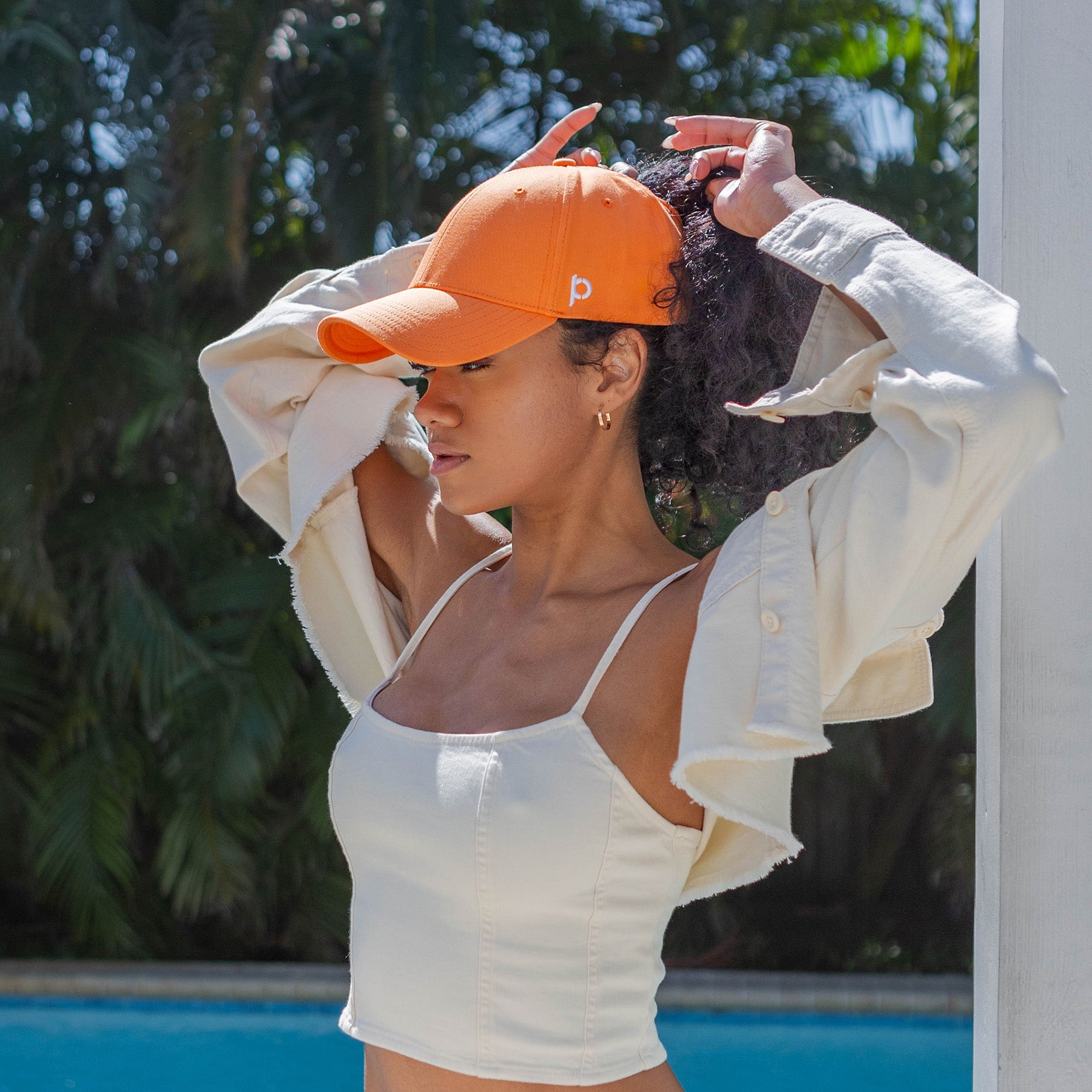 A woman wearing a vibrant orange baseball cap adjusts her cap near a pool setting with tropical foliage in the background, showcasing a stylish sporty look.